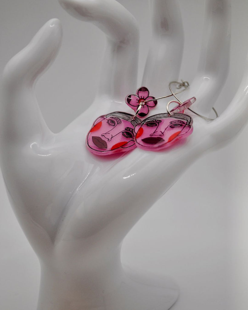 Foto van paar kleine oorbelletjes, gepresenteerd op een witte hand, tegen een witte achtergrond. De oorbellen zijn 2 roze gezichtjes met bloemen details. De kleuren zijn roze, oranje en donkerrood