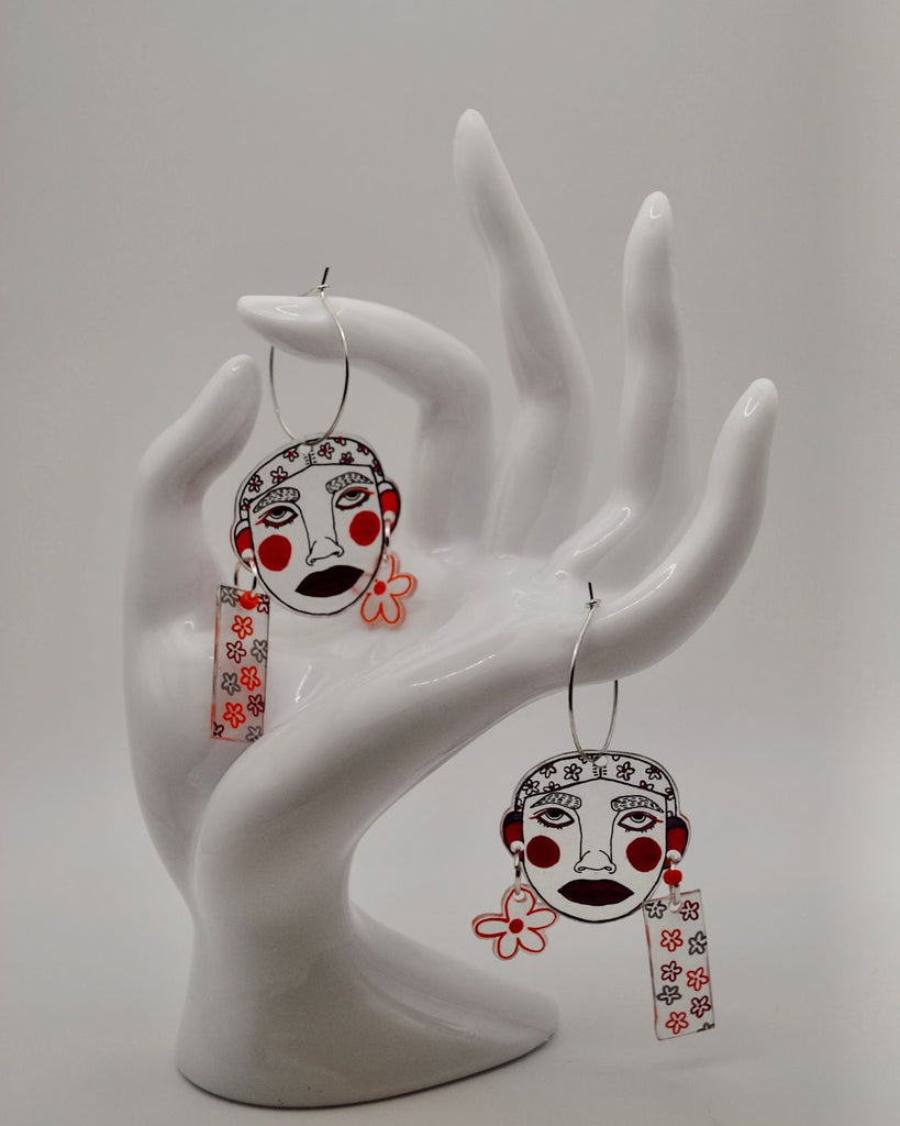 Foto van hangende oorbellen tegen een witte achtergrond, gepresenteerd op een wit hand. De oorbellen zijn 2 gezichtjes met blozende wangetjes. De gezichtjes hangen aan een klein oorringetje én ze hebben zelf ook 2 mini oorbelletjes met bloemetjes 