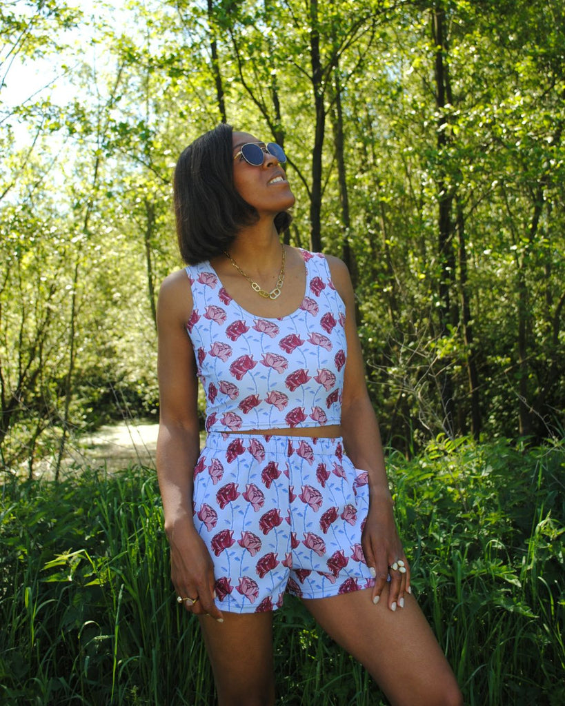 Het vrouwelijke model draagt een crop top en matching short. De set is helderblauw met bloemenprint in roze, bordeaux en blauw. De short heeft zakken.