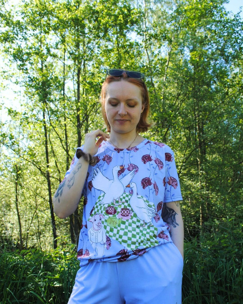 Het model kijkt naar beneden. Ze draagt een comfy blauwe t-shirt in zomers blauw met een uniek print. De prints toont bloemetjes, een picknick tafereel met ganzen en een biggetje.