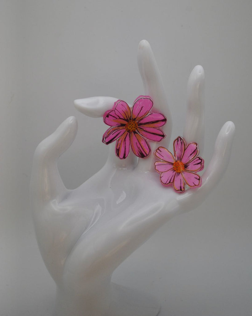 Foto van een paar matching bloemen ringen. De bloemen zijn roze met oranje en gele details. 1 van de ringen is wat groter dan de andere. De ringen worden op deze foto gepresenteerd aan een witte hand en tegen een witte achtergrond.