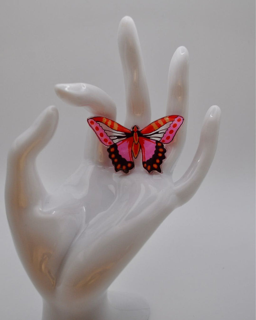 Foto van dichterbij van een unieke statement ring van een vlinder . De ring is met de hand getekend. De vlinder is roze met oranje en donkerrood met een paar doorschijnende details om de vleugels van een vlinder te imiteren. Op deze foto wordt de ring gepresenteerd aan een witte hand en tegen een witte achtergrond. 