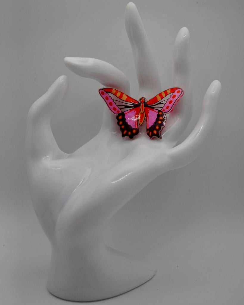 Foto van een unieke vlinder statement ring. De ring is met de hand getekend. De vlinder is roze met oranje en donkerrood met een paar doorschijnende details om de vleugels van een vlinder te imiteren. Op deze foto wordt de ring gepresenteerd aan een witte hand en tegen een witte achtergrond. 
